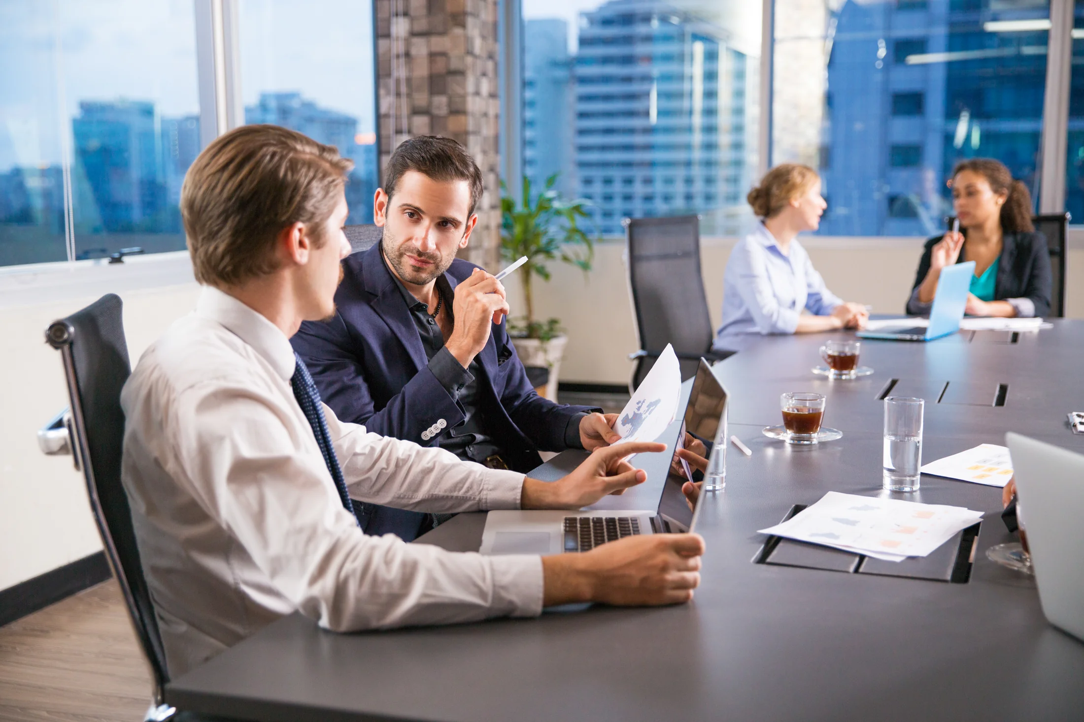 Two pairs of colleagues discussing work at a long wooden table in a bright office