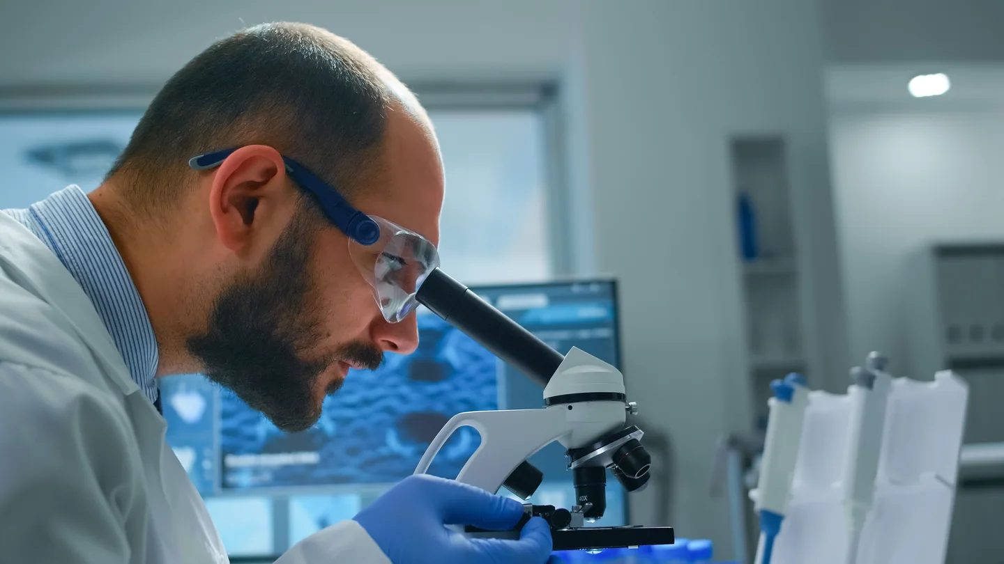 Stock photograph of a laboratory scientist in safety glasses examining a sample through a microscope